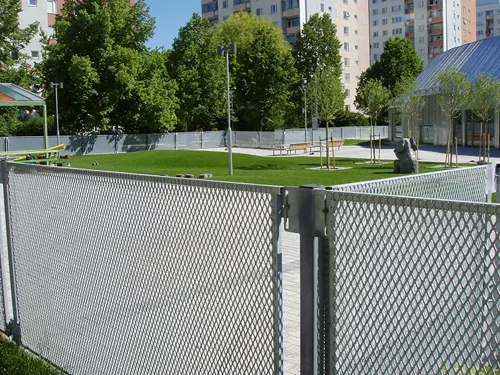 Staket av plattvalsad sträckmetall i ett stadsnära parkområde, med galvaniserad yta och modern nätstruktur, omgärdar en grön yta med bänkar, konstverk och nyplanterade träd, omgiven av höghus och grönska i bakgrunden